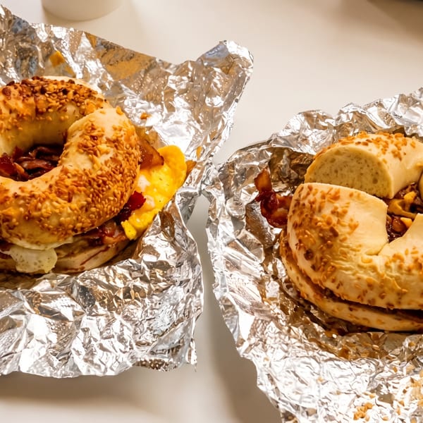 Two bagels with egg, cheese, and bacon, wrapped in aluminum foil on a white surface.